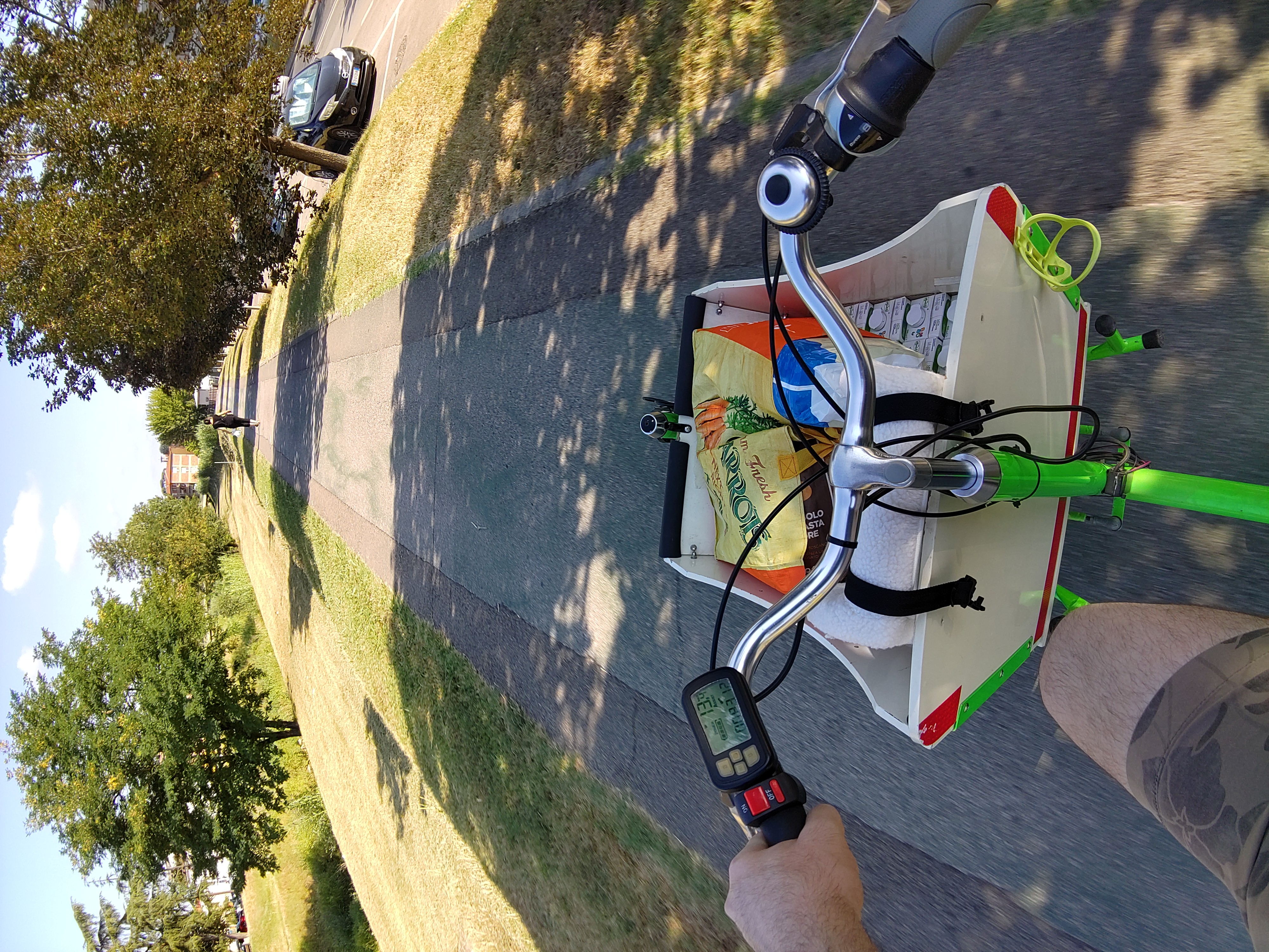 A green front loader cargo bike, with a white loading crane, full of groceries bag and milk boxes partially covering a child seat, seen from the saddle, coasting along a cycle path between a road and a small river.